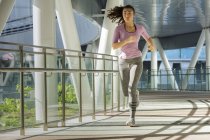 A young asian woman is jogging through the city of Singapore in the early morning. She passes a section of steel and glass architecture. — Stock Photo
