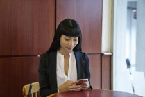 Молодая привлекательная азиатская предпринимательница, использующая смартфон в современном офисе — стоковое фото