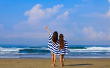 Dos amigas con el pelo largo están de pie en una playa envuelta en una toalla de rayas azules y blancas disfrutando de la vista al mar . - foto de stock