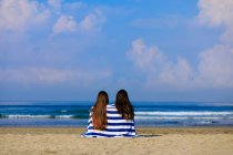 Duas amigas com cabelos longos estão sentadas em uma praia cercada de ablue e toalha listrada branca desfrutando de vista mar teh. — Fotografia de Stock