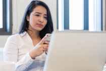 Заинтересованная молодая женщина за ноутбуком, держит мобильный телефон в современном офисе — стоковое фото