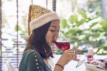 Happy asian woman drinking wine in christmas hat — Stock Photo