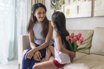 Giovane madre asiatica con figlia carina a casa, ragazza che tiene i fiori nel bouquet dietro la schiena — Foto stock