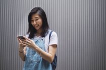 Молодой студент азиатского колледжа, использующий смартфон против серой стены — стоковое фото