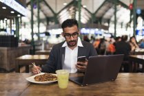 Homem de negócios indiano bonito usando smartphone e comer no café — Fotografia de Stock