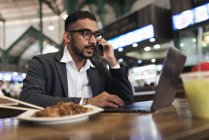 Hombre de negocios indio guapo usando teléfono inteligente y comer en la cafetería - foto de stock