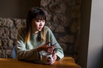 Молодая случайная азиатская девушка, использующая смартфон в кафе — стоковое фото