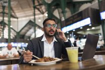 Hombre de negocios indio guapo usando teléfono inteligente y comer en la cafetería - foto de stock