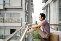 Side view of adult asian man drinking coffee on balcony at home — Stock Photo