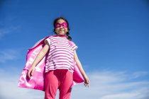 Nettes kleines Superheldenmädchen in Maske und Mantel posiert gegen den Himmel — Stockfoto