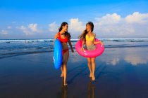 Dos chicas guapas con flotadores camino a las olas del océano. Una chica se da la vuelta sonriendo . - foto de stock