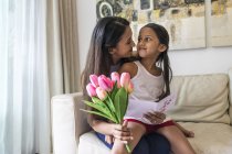 Junge asiatische Mutter mit süßen Tochter umarmt zu Hause mit Blumen und Vorlage — Stockfoto