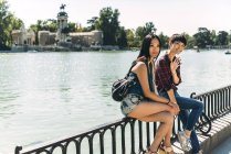 Japanese and Chinese female friends travellers sitting on handrail and looking at camera in Retiro park, Madrid, Spain. — Stock Photo