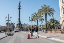 Young tourist couple crossing the road at the Columbus monument with a suitcase, spain — Stock Photo