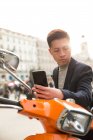 Un joven chino casual con un teléfono inteligente. sentado en una moto en Puerta del Sol, Madrid, España - foto de stock