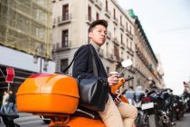 Jeune Chinois décontracté avec un téléphone intelligent assis sur une moto à Puerta del Sol, Madrid, Espagne — Photo de stock