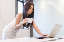 Jovem mulher verificando seu laptop com xícara de café no escritório moderno — Fotografia de Stock