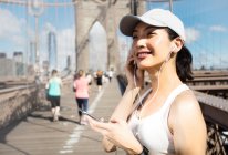 Довольно азиатская бегунья тренируется на открытом воздухе, работая на Бруклинском мосту с Манхэттеном, Нью-Йорк на заднем плане . — стоковое фото