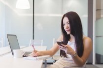 Junge Frau macht sich in modernem Büro Notizen mit Handy in der Hand — Stockfoto
