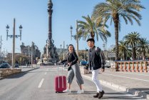 Jeune couple touristique traversant la route au monument de Colomb avec une valise — Photo de stock