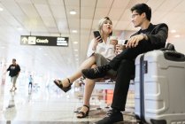Young asian couple of businesspeople in airport with smartphone and drink — Stock Photo