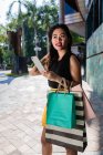 Sonriendo mujer asiática con bolsas de compras usando teléfono inteligente - foto de stock