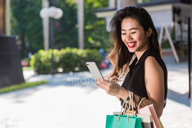 Усміхнена азіатська жінка з сумками за допомогою смартфона — стокове фото