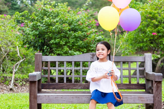 Kleines Mädchen auf Parkbank mit Luftballons. — Stockfoto