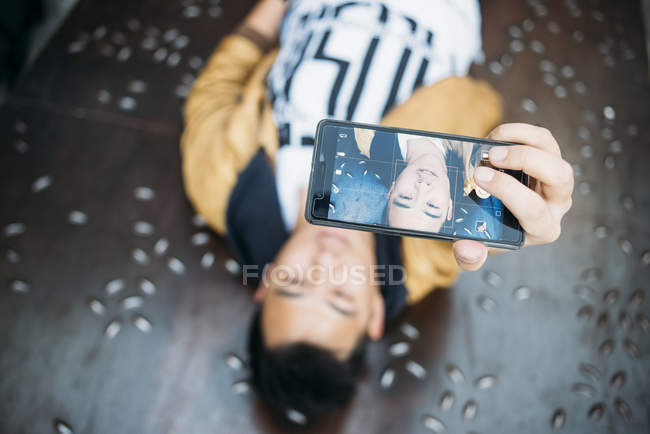 Joven asiático macho acostado en suelo y tomando selfie - foto de stock