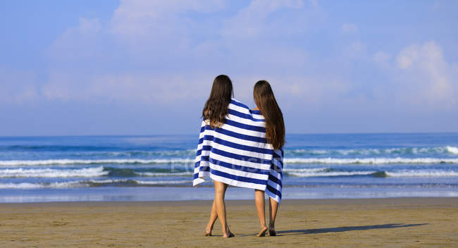 Duas amigas com cabelos longos estão em pé em uma praia envolta em uma toalha listrada azul e branca desfrutando da vista do mar . — Fotografia de Stock