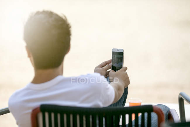 Junger Mann sitzt draußen an einem Strand und blickt mit Smartphone in der Hand in die Ferne — Stockfoto