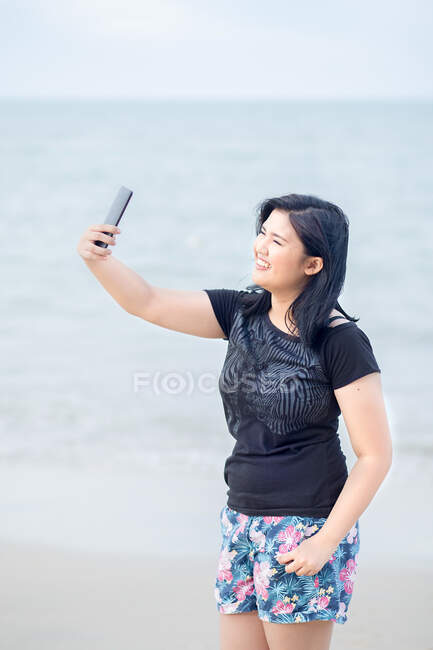 Adolescente con teléfono inteligente tomando selfie por la playa. - foto de stock