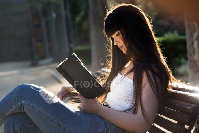 Junge eurasische Frau liest ein Buch auf Bank im Park — Stockfoto