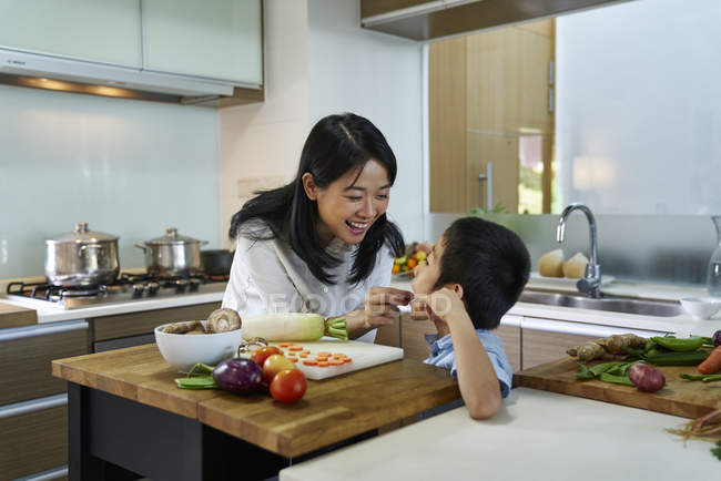 Happy young asian mother feeding son at kitchen — Stock Photo