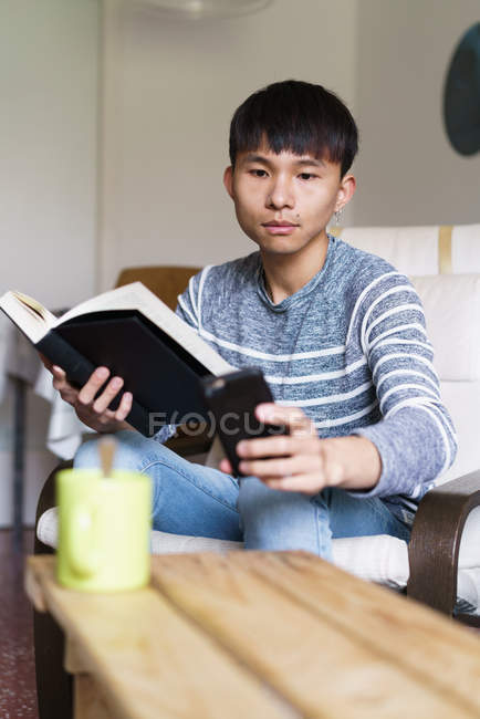 Азиатский мужчина дома с книгой, заглядывающей в его телефон — стоковое фото