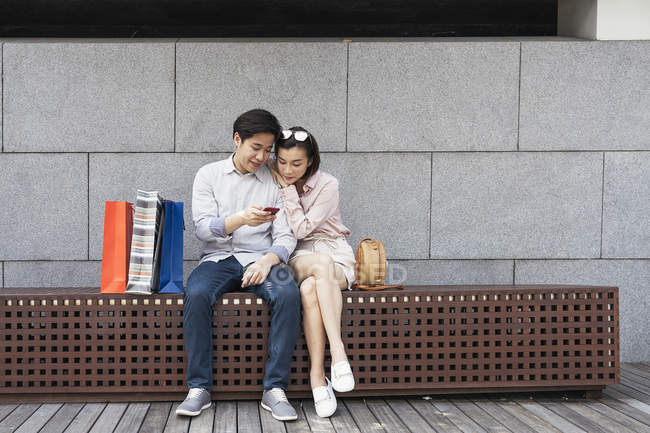 Joven asiático pareja usando smartphone juntos en banco - foto de stock