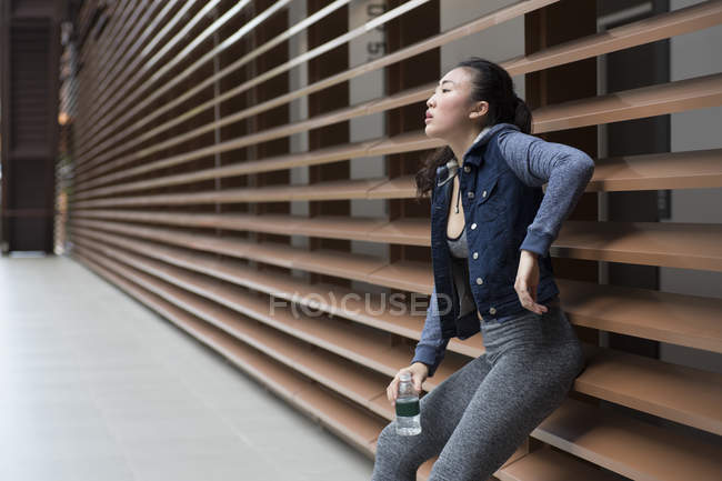 Uma jovem asiática está descansando em uma parede depois de seu treino correndo através de sua vizinhança. Ela está segurando uma garrafa de água . — Fotografia de Stock