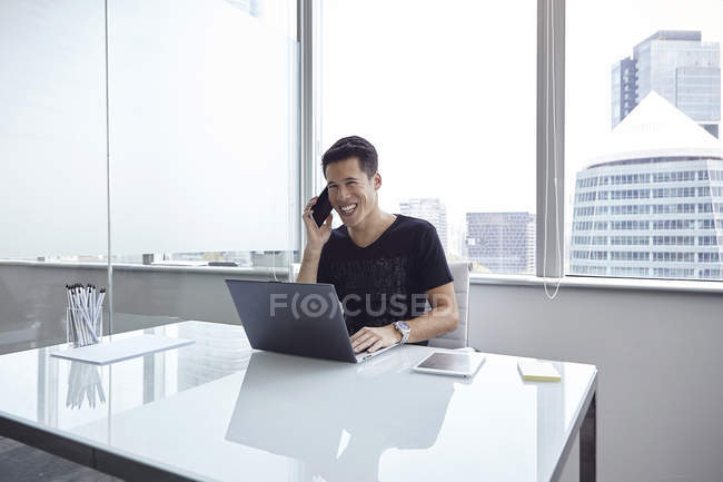 Handsome asian businessman talking by phone and using laptop at home — Stock Photo