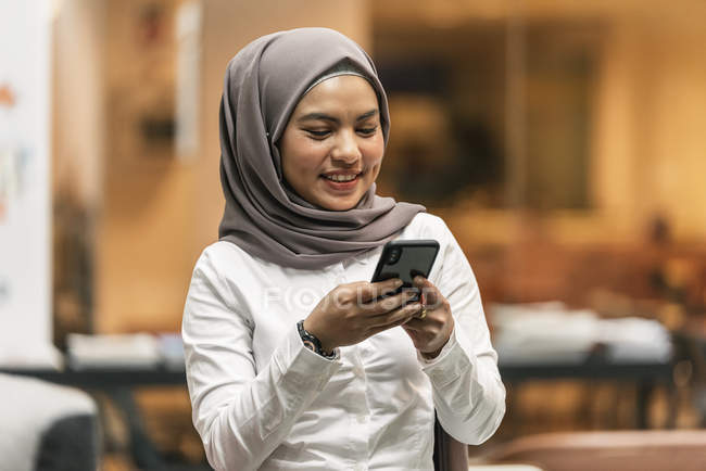 Jovem asiático mulher de negócios no hijab usando smartphone no escritório moderno — Fotografia de Stock