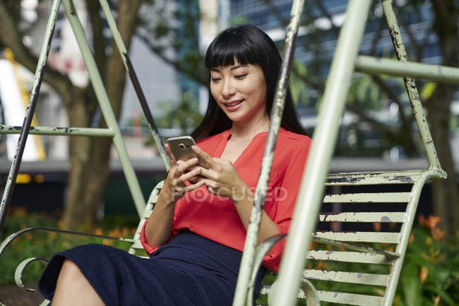 Jeune femme utilisant un téléphone portable sur swig dans le parc, Singapour — Photo de stock