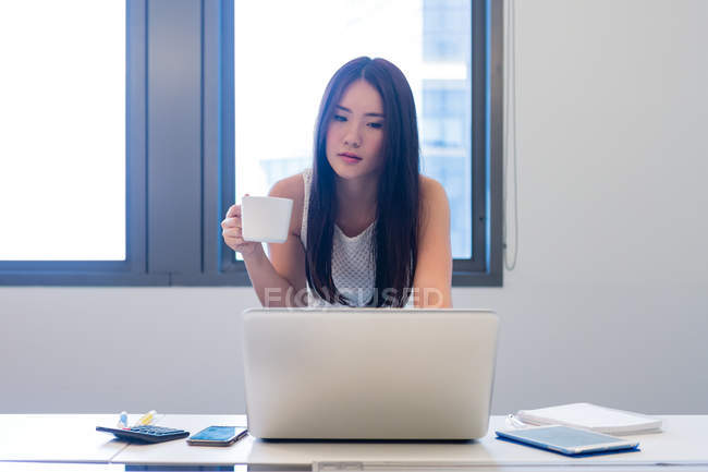 Mujer joven sosteniendo coffeecup y revisando su computadora portátil . - foto de stock