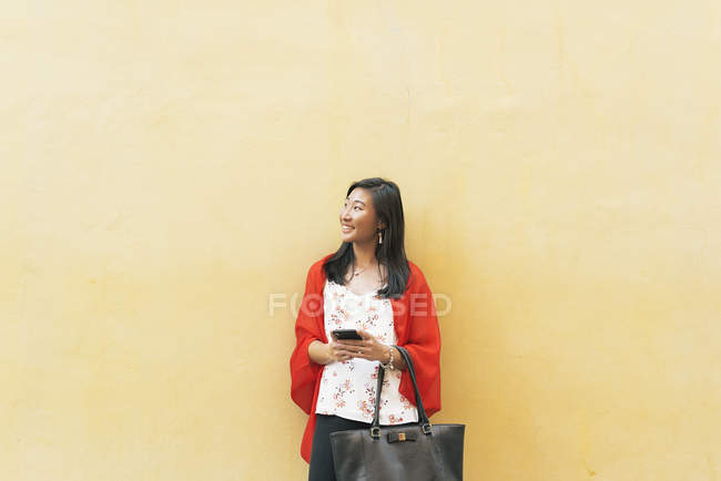 Giovane donna asiatica posa contro muro giallo con smartphone — Foto stock