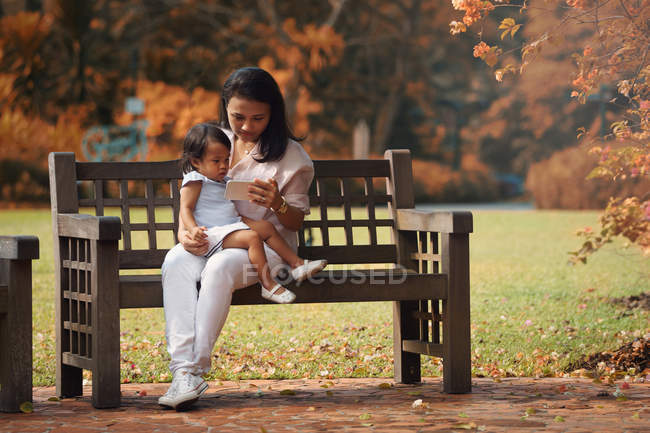 Cute asian mother and daughter using smartphone on bench in park — Stock Photo