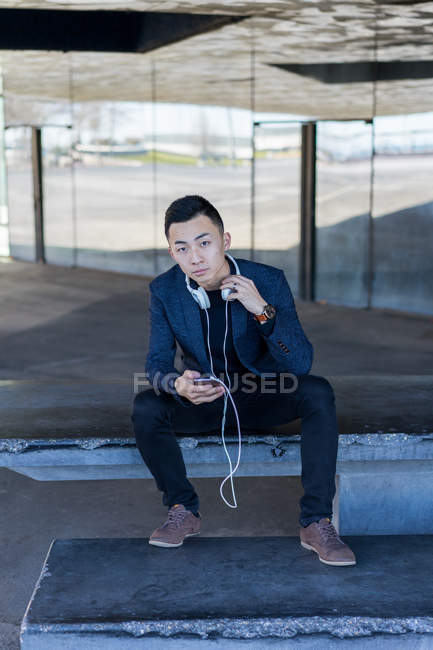 Young asian man with smartphone and headset sitting on street — Stock Photo