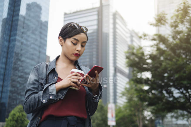 Jeune femme malaise singapourienne dans un cadre urbain avec son smartphone et une tasse de café dans les rues . — Photo de stock