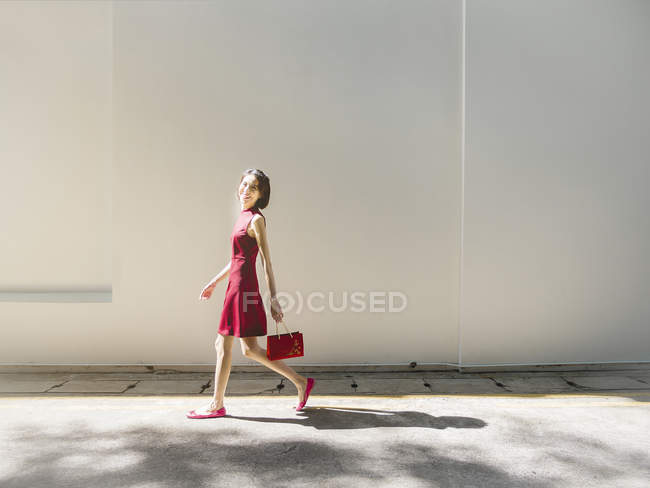 Mulher chinesa andando contra a parede branca com bolsa — Fotografia de Stock