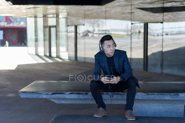 Молодий азіатський чоловік сидить у навушниках на вулиці — стокове фото