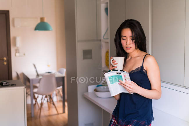 Joven mujer china leyendo una revista con una taza de café en casa - foto de stock