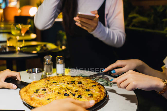 Imagen recortada de amigos cortando pizza en un cómodo bar - foto de stock