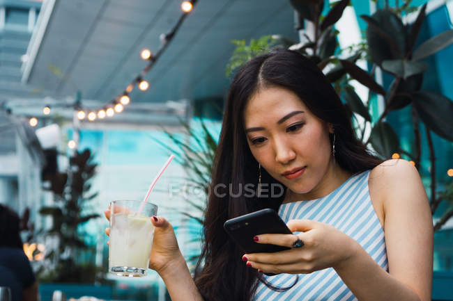Молодая азиатка, использующая смартфон и имеющая напиток — стоковое фото